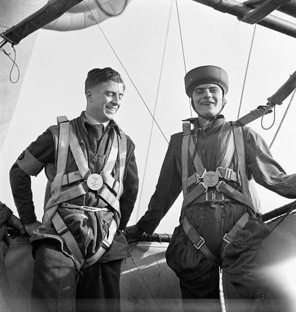 Black and white photograph. Two men in military uniforms and parachutes lean on a rail, laughing.